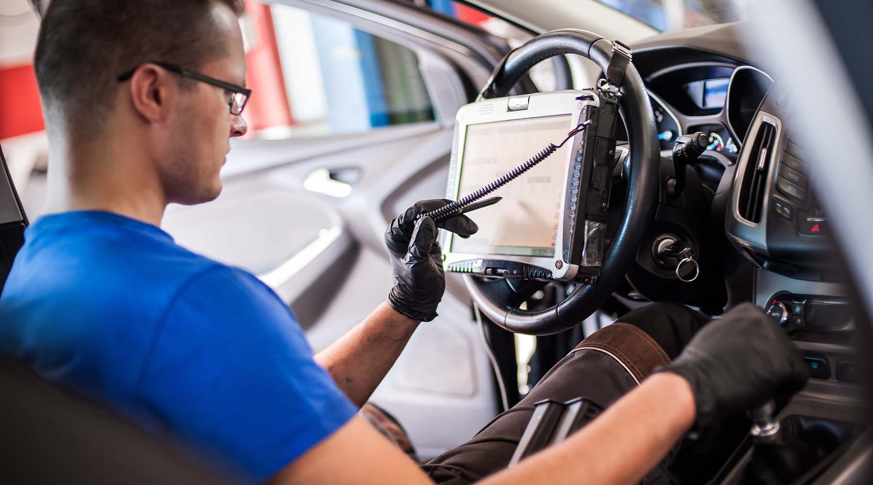 Kfz-Mechatroniker beim Abmessen elektronischer Sicherheitsprüfungen und das Erfassen elektronischer Messwerte an einem Fahrzeug im Innenraum.