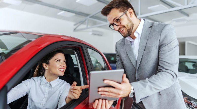 Verkaufsberater Gebrauchtwagen mit einer Kundin in einer Autogalerie die in einem Fahrzeug sitzt und vom Händler am Tablet etwas gezeigt bekommt.