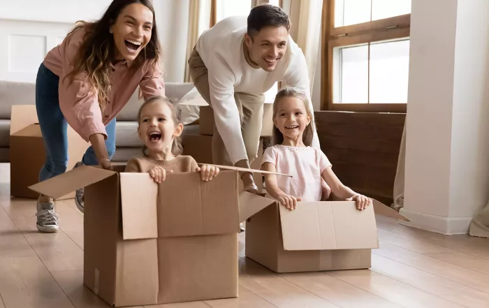 Glückliche Eltern, die mit Kindern im Wohnzimmer der neuen Wohnung spielen, zwei süße kleine Töchter, die in Pappkartons sitzen, lachende Mutter und Vater schieben, Familie feiert den Umzugstag