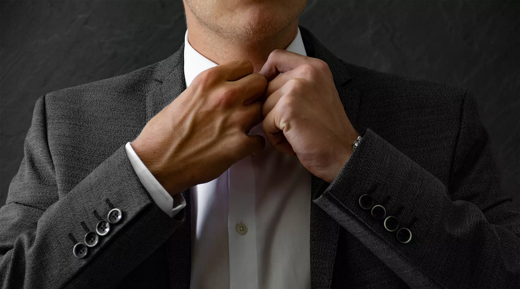 Mann der sich im Anzug den Kragenknopf seines weissen Hemd zuknüpft.